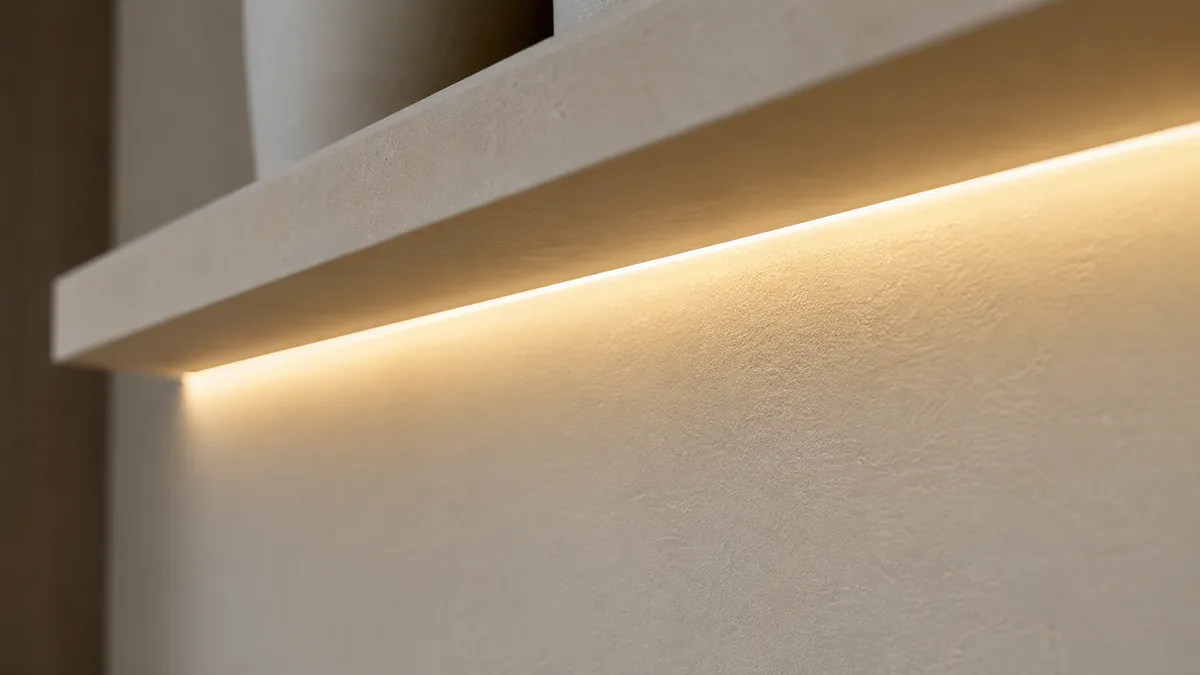 棚の手前に隠したライン照明が、LEDの粒を見せずに一本の連続した暖色の帯として壁を照らす接写。拡散で面光源に近づけた例